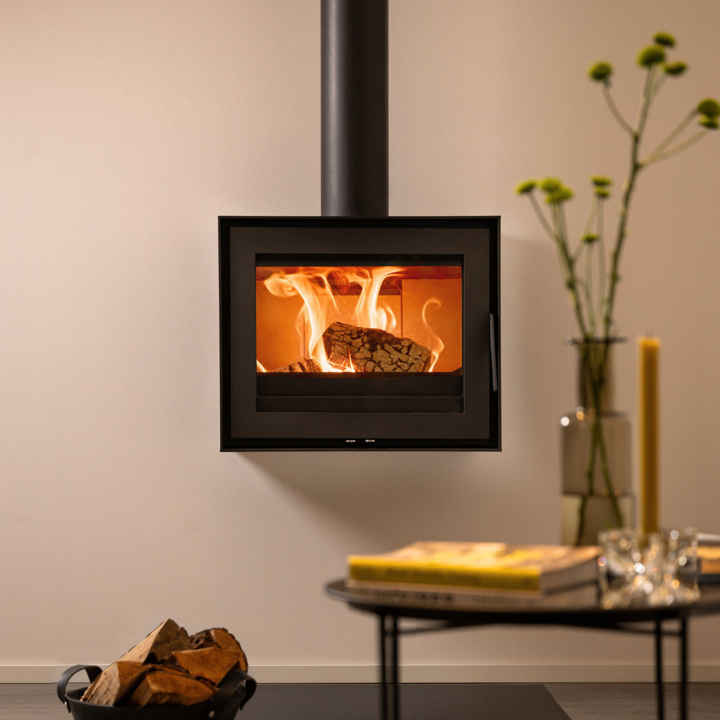 Wanders WAN-2075 Black Edition Low, moderne haard met brandend hout, naast een tafel en bloemen.
