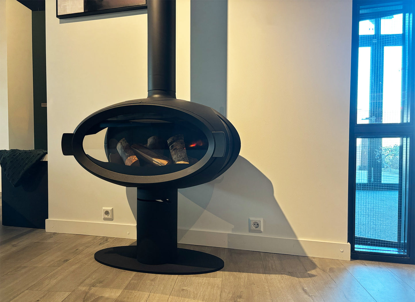 Wanders Mata Round Base, een moderne zwarte houtkachel met een ovale glazen deur.