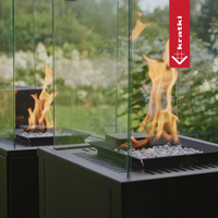 Kratki Patio Mini tuinhaard met vlammen, glazen scherm en witte stenen in een groene buitenomgeving.