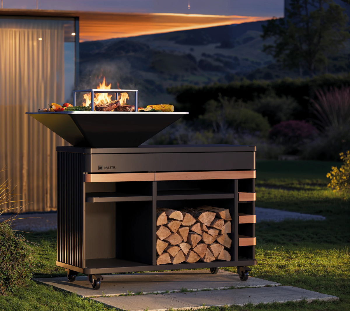 Balstil Big Chef Grill op mobiele buitenkeuken met vlammen, gegrild eten en houtopslag in tuin.
